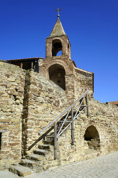 A stone staircase with a wooden handrail ascends the rugged masonry walls toward a pointed brick belfry crowned with a Christian cross at the historic Davit Gareja monastery complex in eastern Georgia