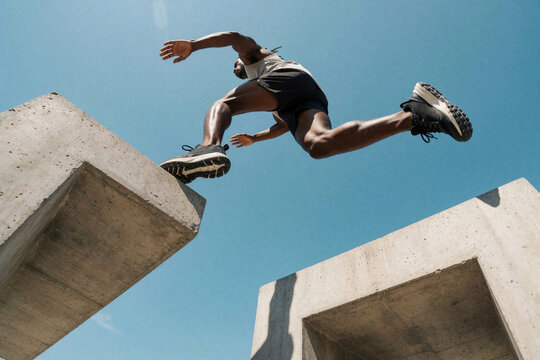 Athletic man jumps between concrete platforms under blue sky. Fit person performs parkour move between urban structures. High action shot shows extreme sport training, jumping over gap, outdoor