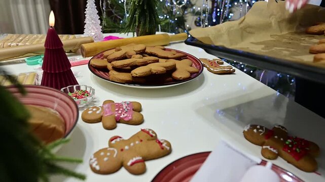 A woman bakes gingerbread men in a kitchen. She places the cookies on a plate while a Christmas tree and garland are seen in the background. The scene shows the fun of holiday baking.