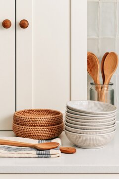 Vertical still life of stacked ceramic bowls, woven rattan baskets, wooden utensils and folded kitchen towel in a warm neutral kitchen, with copy space