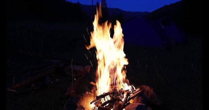 Bright campfire burning with tall vibrant flames near a camping tent under a dark twilight mountain sky.