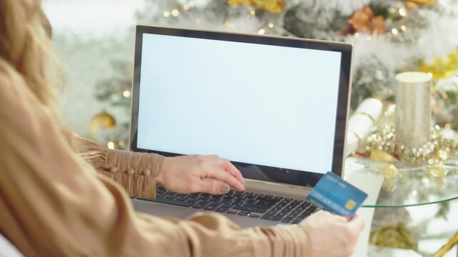 A womans hands type on laptop, holding credit card ready for purchase, set against blurred background of glowing Christmas tree. This captures essence of convenient holiday online shopping.