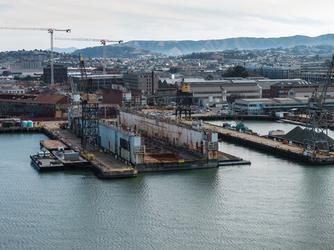 Aerial perspective displays a floating drydock, heavy cranes, and industrial waterfront infrastructure along Pier 70 in San Francisco, California, with distant city hills under a cloudy sky.