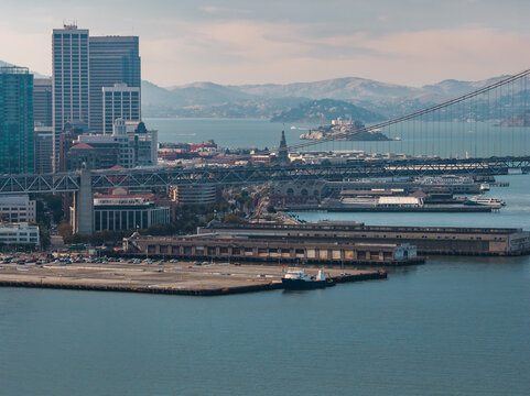 Aerial view captures the San Francisco embarcadero waterfront, showing piers, city buildings, the Bay Bridge span, and Alcatraz Island across the water in California.