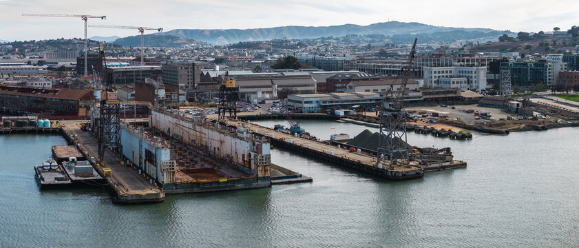 Drydock and maritime shipyard facilities at Pier 70 in San Francisco are viewed across the water, showing industrial cranes, docks, warehouses, and rolling coastal hills under daylight.
