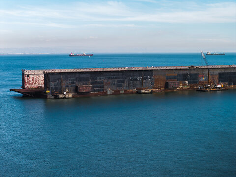 Floating drydock structure rests in the coastal waters of San Francisco Bay, with cargo vessels navigating across the distant marine horizon under clear daytime skies.