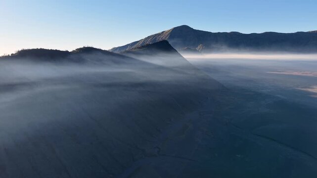 Mist wraps around volcanic slopes in soft, flowing waves. Mountains stand tall near Mount Bromo