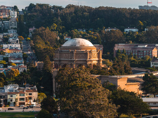 Aerial view shows the Palace of Fine Arts rotunda in San Francisco surrounded by trees and residential buildings, with Sutro Tower visible on the distant horizon during warm afternoon light. © Aerial Film Studio
