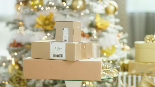 A close-up showcases stack of three cardboard boxes on clear glass table. In softly blurred background, festive Christmas tree, adorned with lights, golden ornaments creates cheerful holiday scene.