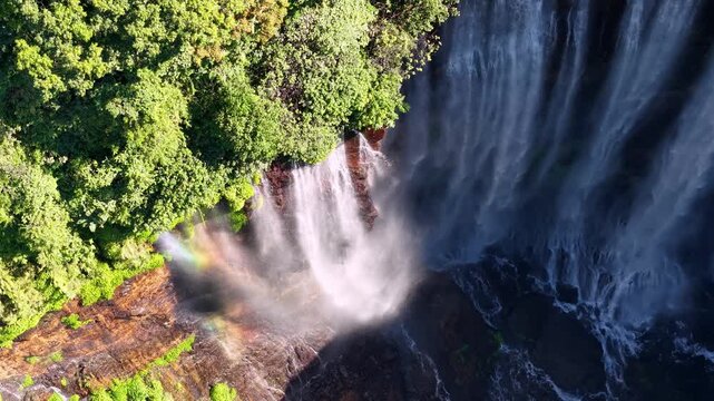 Aerial drone view of Tumpak Sewu waterfall showing lush cliffs, cascading water, misty spray and subtle rainbow
