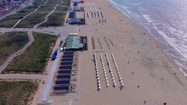 Aerial drone clip of sandy beach coastline, North Sea waves, beach cabins, resort pavilions, and coastal sand dunes in Katwijk aan Zee, South Holland, Netherlands on bright summer day