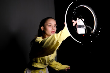Fototapeta premium Young female content creator recording a social media video with smartphone mounted on a ring light, looking into camera in a dark home studio for livestreaming and vlogging