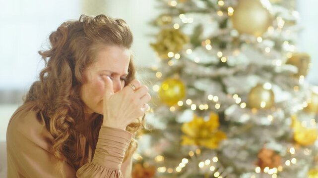 A woman with curly brown hair, wearing tan blouse, sits in profile, hand touching face with distressed expression. Before blurred Christmas tree, soft lighting reflects sadness and holiday blues.