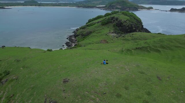 Solitary Couple On Grassy Coastal Hill Overlooking Ocean And Rocky Islet, Drone Panorama Captures Sweeping Green Slopes, Gentle Waves On Shore, Quiet Relaxation And Exploration