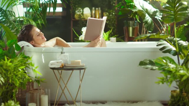 A smiling woman with a bun relaxes in a white bathtub, reading a book. Bathed in natural light, the plant-filled bath creates a peaceful, spa-like setting, perfect for self-care & literary escape.