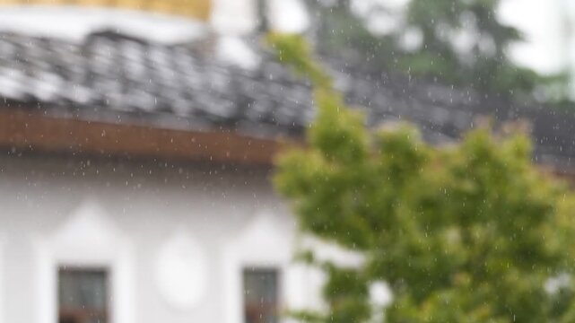 Light rain drizzles against building and tree foliage, seasonal weather change from summer to autumn, many blurred raindrops falling down outside at residential house backyard during rainfall shower.