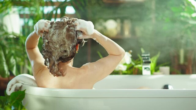 A woman from the back indulges in a relaxing home spa, washing her curly hair with foamy shampoo in a white bathtub. The bathroom, bathed in natural light & greenery, suggests tranquility & pampering.
