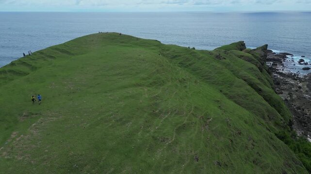 Couple Hiking Along Coastal Ridge Aerial Drone View Showing Two Hikers In Blue And Yellow Jackets Crossing Green Grassy Hill Above Ocean And Rocky Islets, Overcast Sky, Peaceful Travel Mood,Lombok