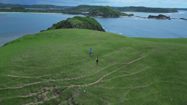 Woman nad Man Walking Across Green Hillside Path, Small Lone Figure Following Worn Trails, Aerial Perspective Highlights Winding Tracks And Rolling Grass, Calm Overcast Light, Bukit Merese, Lombok