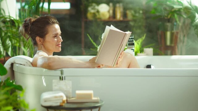 A serene woman with a bun relaxes in a white bathtub, engrossed in a book. Surrounded by lush greenery & soft steam, the scene in bright natural light exudes tranquility & well-being.