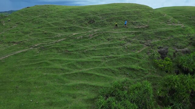 Woman nad Man Walking Across Green Hillside Path, Small Lone Figure Following Worn Trails, Aerial Perspective Highlights Winding Tracks And Rolling Grass, Calm Overcast Light, Bukit Merese, Lombok