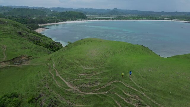 Explorers Standing At Cliff Edge Overlooking Turquoise Bay And Curved Beach, Aerial View Shows Reef Patterns In Shallow Water, Green Hillside Foreground, Contemplative Lookout