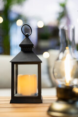 Fototapeta premium Close-up shows a black candle lantern and oil lamp glowing on a wooden rooftop table near comfortable couch seating, creating minimalist balcony ambiance.