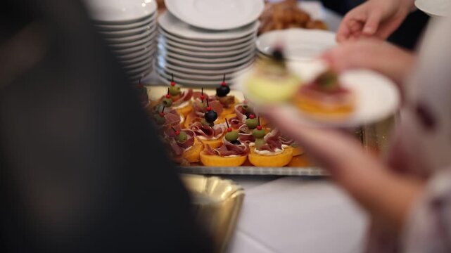 Elegant canapes topped with cured ham, olives, and cocktail picks arranged on a tray at a catered event, with a guest's hand reaching to take one from the buffet table.