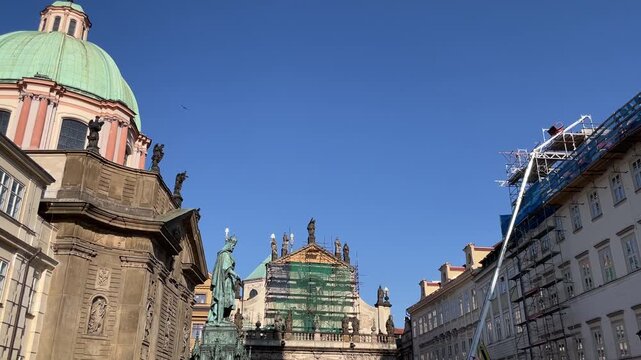 Historic urban streetscape showing pale green copper dome, ornate stone church facades and rooftop statues while central facade is wrapped in green