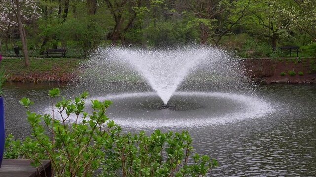 Water fountain spraying droplets in slow motion over a park pond surrounded by lush spring greenery and benches. Concept of relaxation, peaceful outdoor spaces and landscaped gardens.