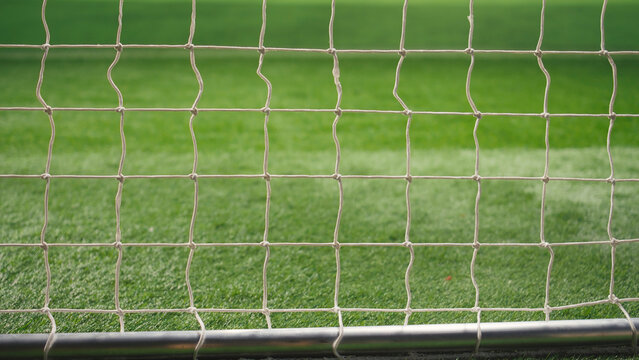 Selective focus shows a side goal post with rope net and knots on a stadium pitch, with an empty football goal and blurred sports arena in the background.