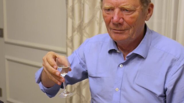 Elderly man wearing light blue button-down shirt holding up small glass of clear liquid and drinking from it inside room with simple wall paneling and light-colored curtain.