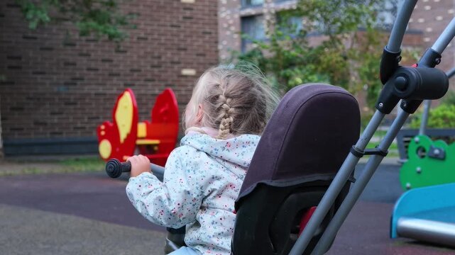 Young blonde girl with braided hair sits in stroller at playground, looking around with slightly teary expression while holding handle, with colorful play equipment in background.
