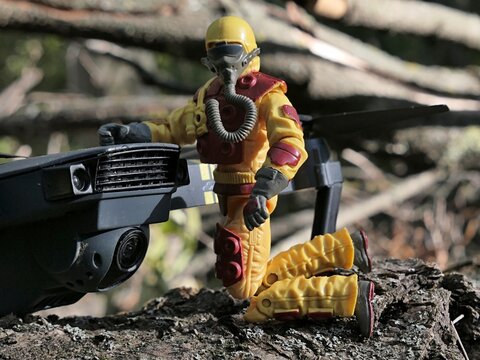Detail of GI JOE action figure of flamethrower man and firefighter Blowtorch, version 2009, dressed in fireproof suit, kneeling on arid soil next to Eachine e58 ultralight quadcopter drone.