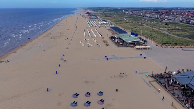 High-angle aerial drone clip of wide sandy beach coast, North Sea waves, beach pavilions, cabins, and coastal town skyline in Katwijk aan Zee, South Holland, Netherlands on bright summer day