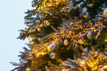 Fototapeta premium Christmas tree decorated with gold and silver ornaments and warm glowing string lights close up, creating festive holiday atmosphere against a soft blue evening background. Street, outside decorations