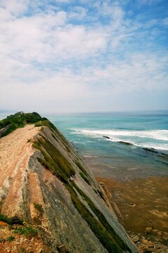 A high angle view of a steep cliff path at Itzurun beach overlooking the sea waves and geological flysch rock formations under a cloudy sky in Zumaia, Spain, representing Basque Country travel.