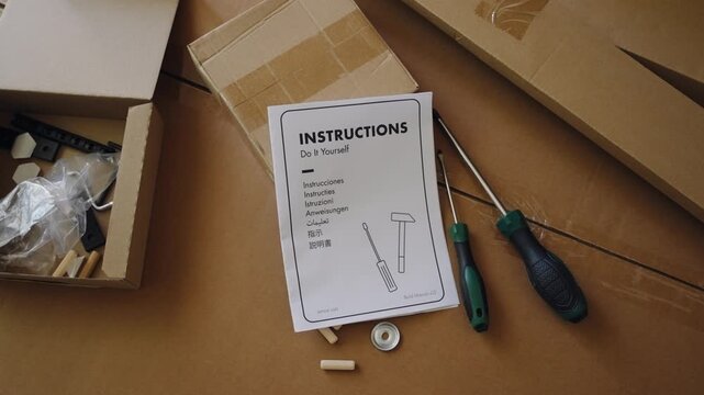 Top view of person dropping a do it yourself instruction manual onto a pile of cardboard boxes, parts, and tools for assembling new furniture in a room with warm natural light