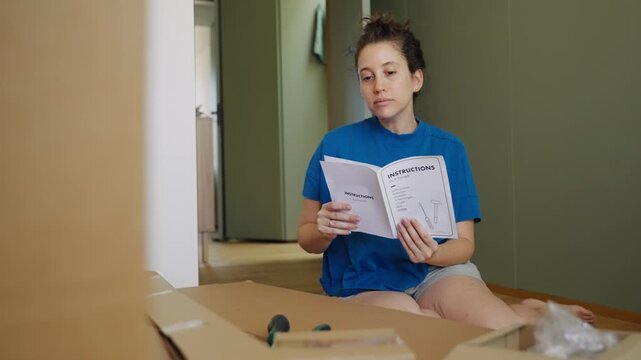 Woman reading instructions for furniture assembly project. Young female home owner holding DIY do it yourself manual before building a furniture. Cardboard boxes and various parts and tools on floor