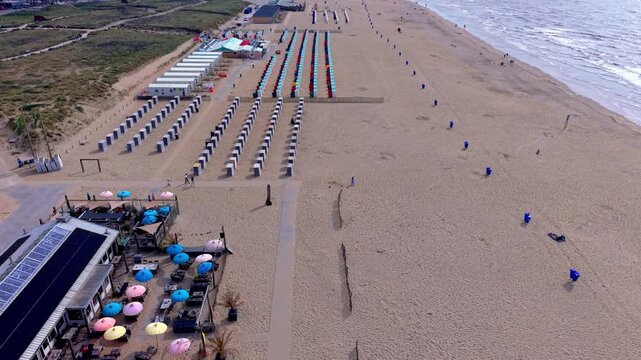 High-angle aerial drone clip of organized beach cabins, colorful umbrellas, beach pavilion, North Sea shoreline, and sandy coast in Katwijk aan Zee, South Holland, Netherlands on sunny summer day