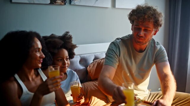 Family clinking orange juice glasses bed. Mother father daughter home breakfast.