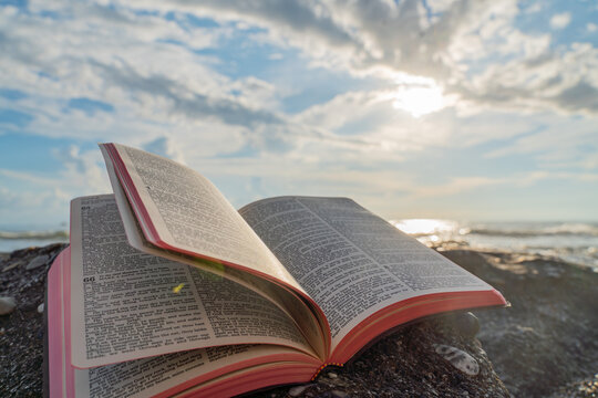 Open Bible Book with Fluttering Pages on Rocky Beach at Sunset