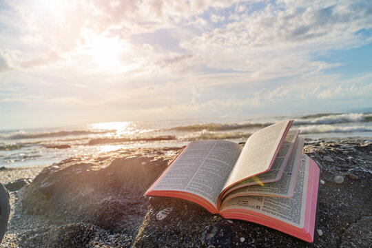 Open Bible Book with Fluttering Pages on Rocky Beach at Sunset