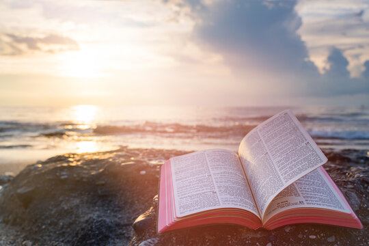 Open Bible Book with Fluttering Pages on Rocky Beach at Sunset