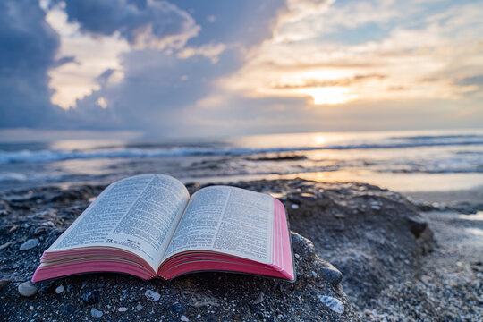 Open Bible Book with Fluttering Pages on Rocky Beach at Sunset