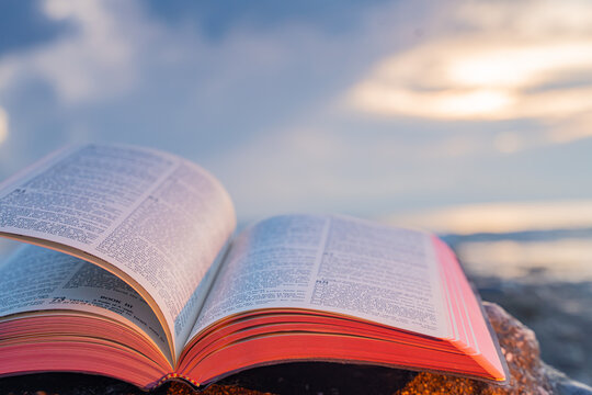 Open Bible Book with Fluttering Pages on Rocky Beach at Sunset