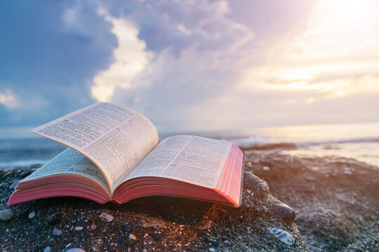 Open Bible Book with Fluttering Pages on Rocky Beach at Sunset
