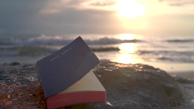 Open Bible Book with Fluttering Pages on Rocky Beach at Sunset