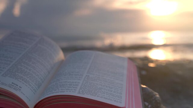 Open Bible Book with Fluttering Pages on Rocky Beach at Sunset