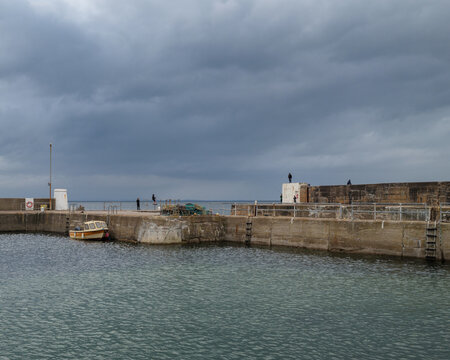 People fishing from the stone harbor walls at Hopeman Harbour in Moray Scotland under a dark and cloudy sky during the afternoon